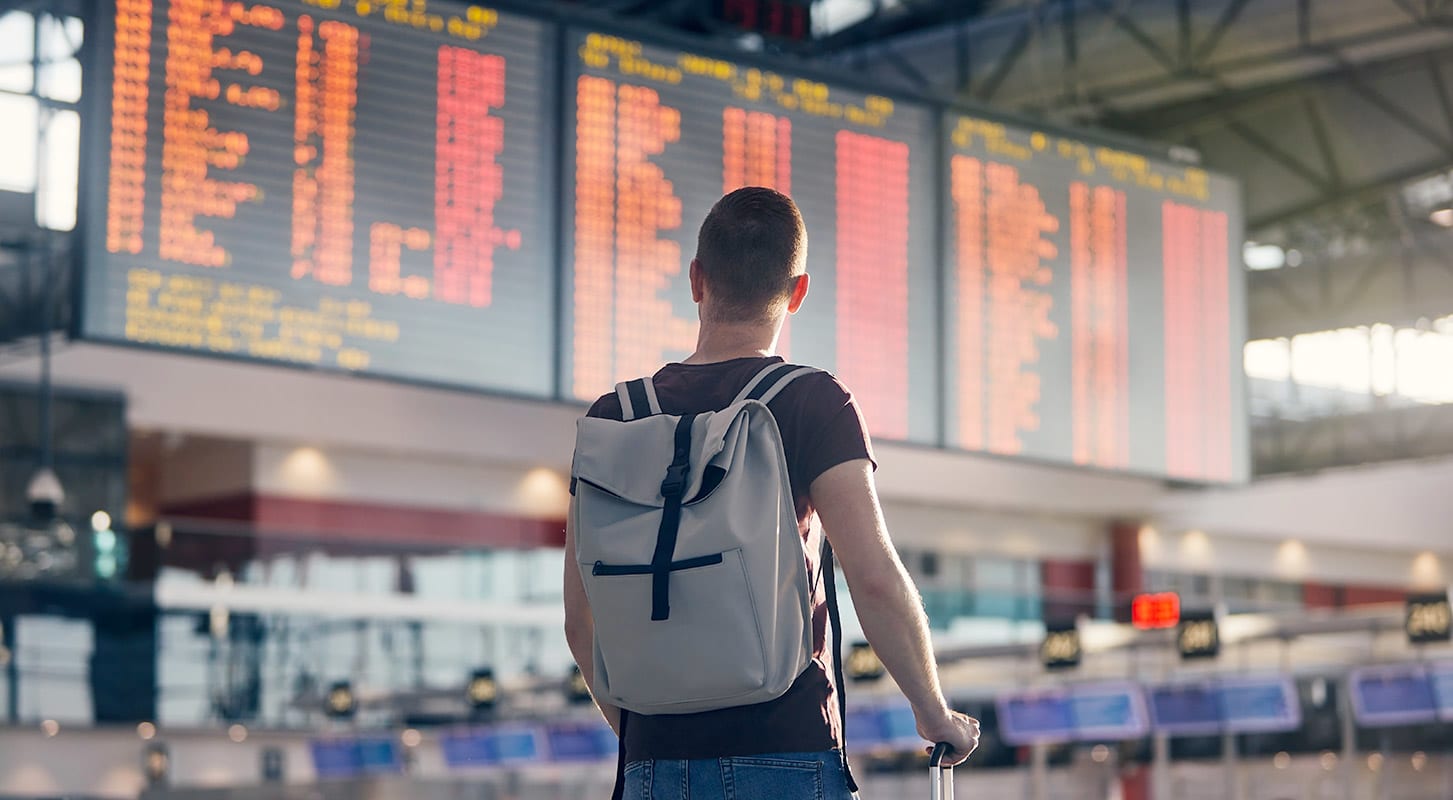 A man stands at the airport with a backpack and luggage, preparing for his journey as he looks at the flight timetable.